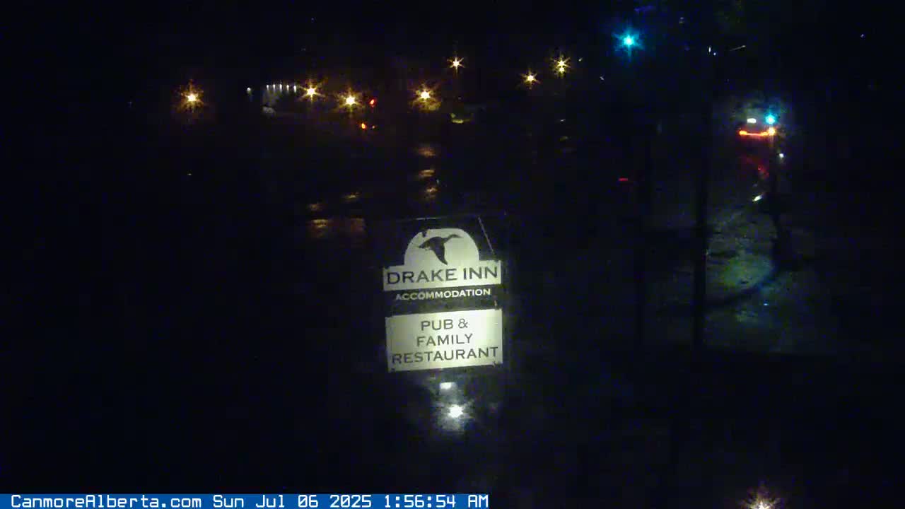 A dark, nighttime scene shows a Drake Inn sign illuminated in front of a wet, dark road with distant lights and vehicles.
