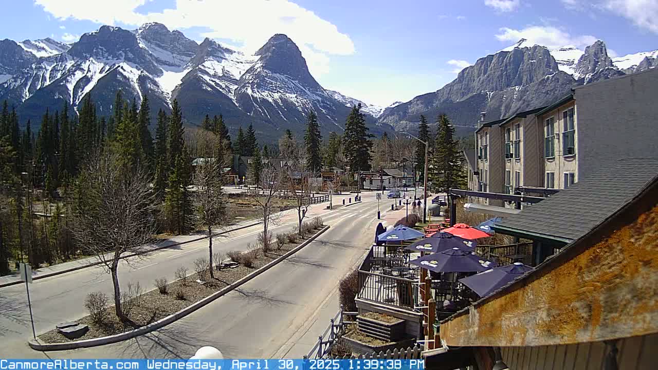 A mostly sunny day reveals a mountain town street scene with snow-capped mountains in the background, featuring a road, some buildings, trees, and an outdoor patio area with umbrellas.