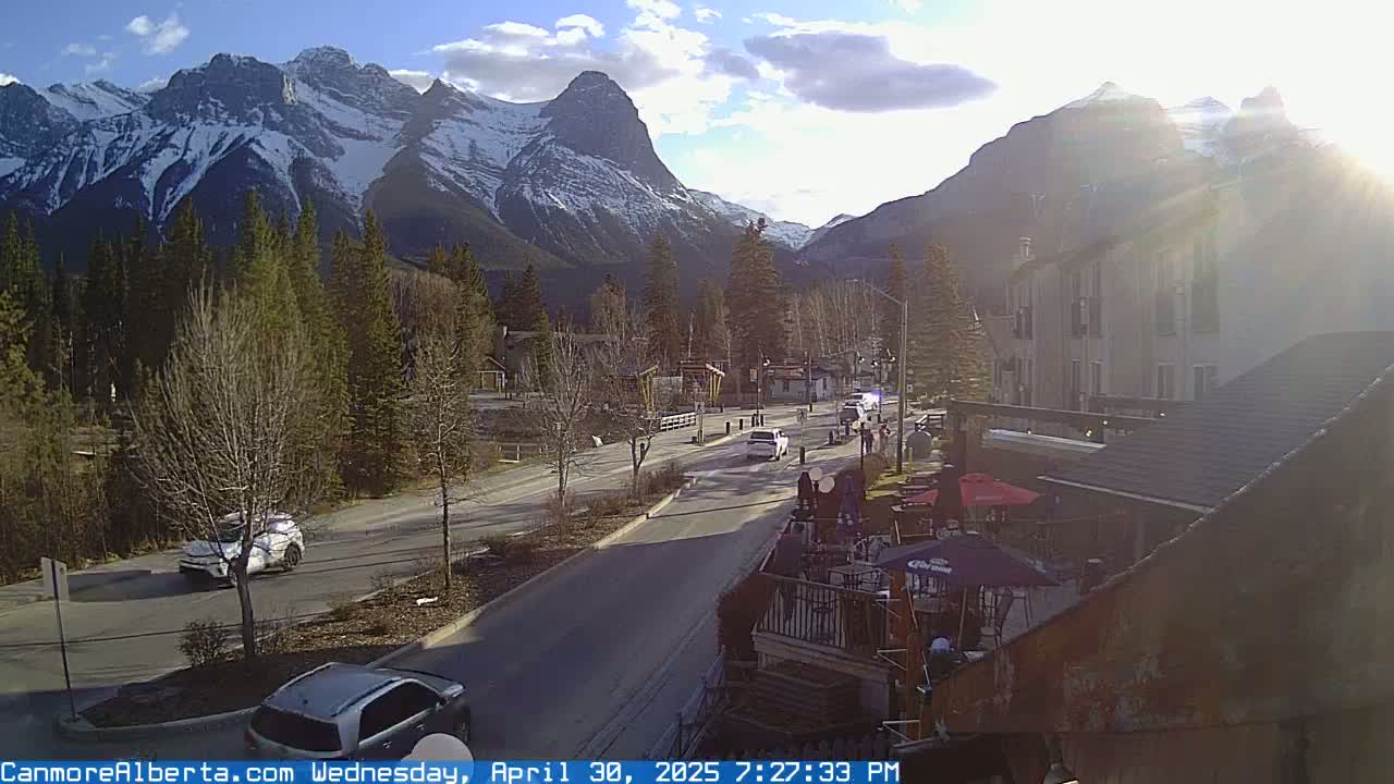 A sunny afternoon view of a mountain town street, with snow-capped mountains in the background and cars driving on the road.