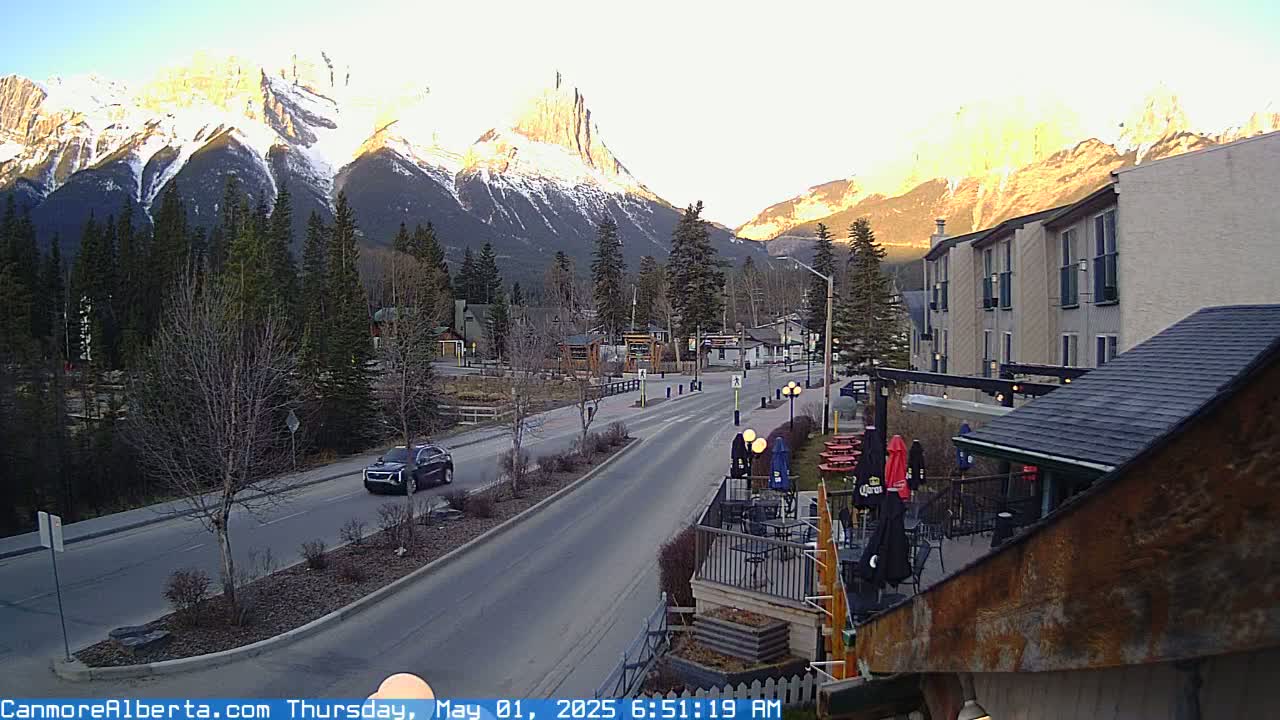 A mountain town street scene at sunrise, with snow-capped mountains in the background, a road with a few cars, and some buildings including a hotel and patio area.