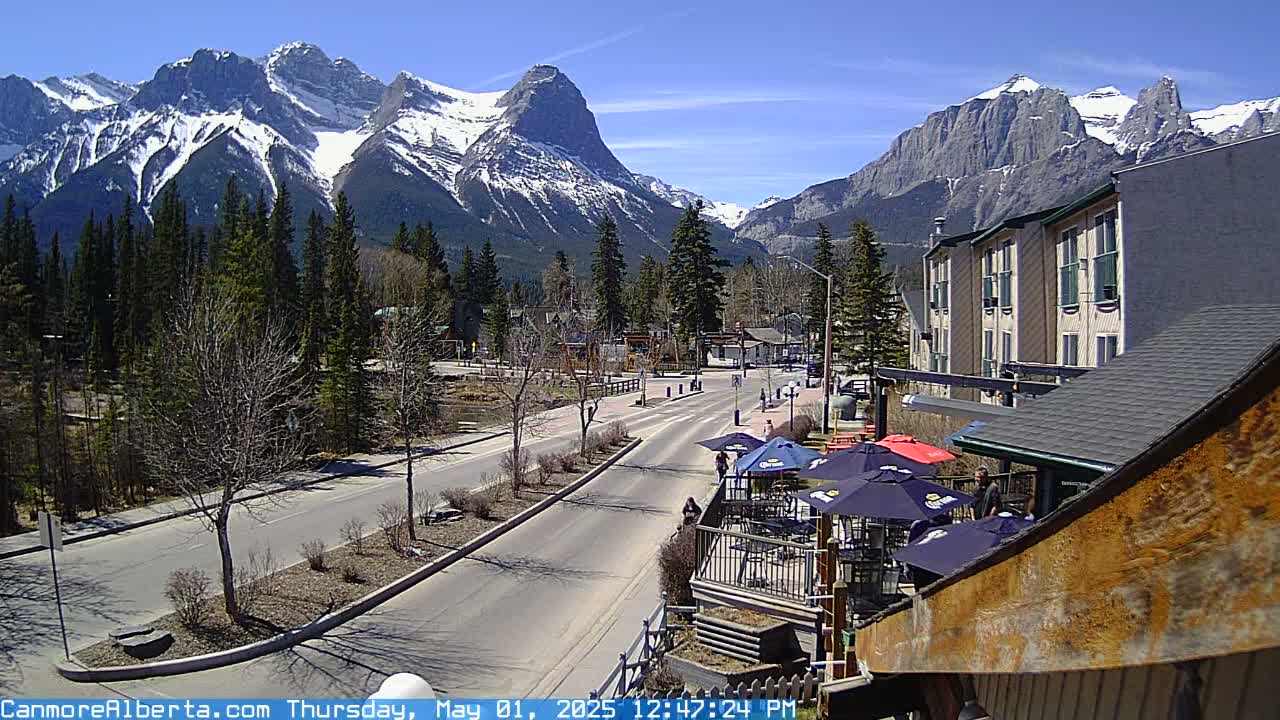 A sunny day reveals a mountain range capped with snow, overlooking a town with a road leading to a patio with umbrellas.