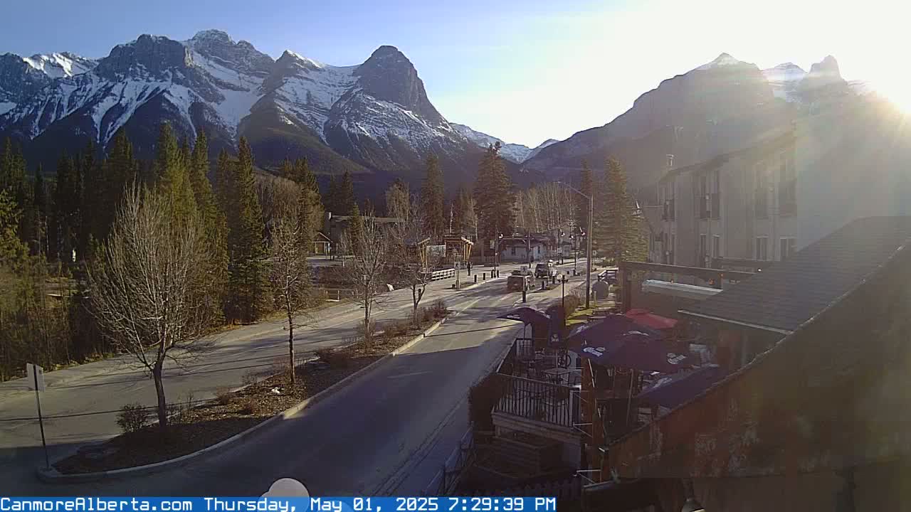 A sunny day reveals snow-capped mountains behind a mountain town with a roadway, trees, and buildings, including a patio area with umbrellas.