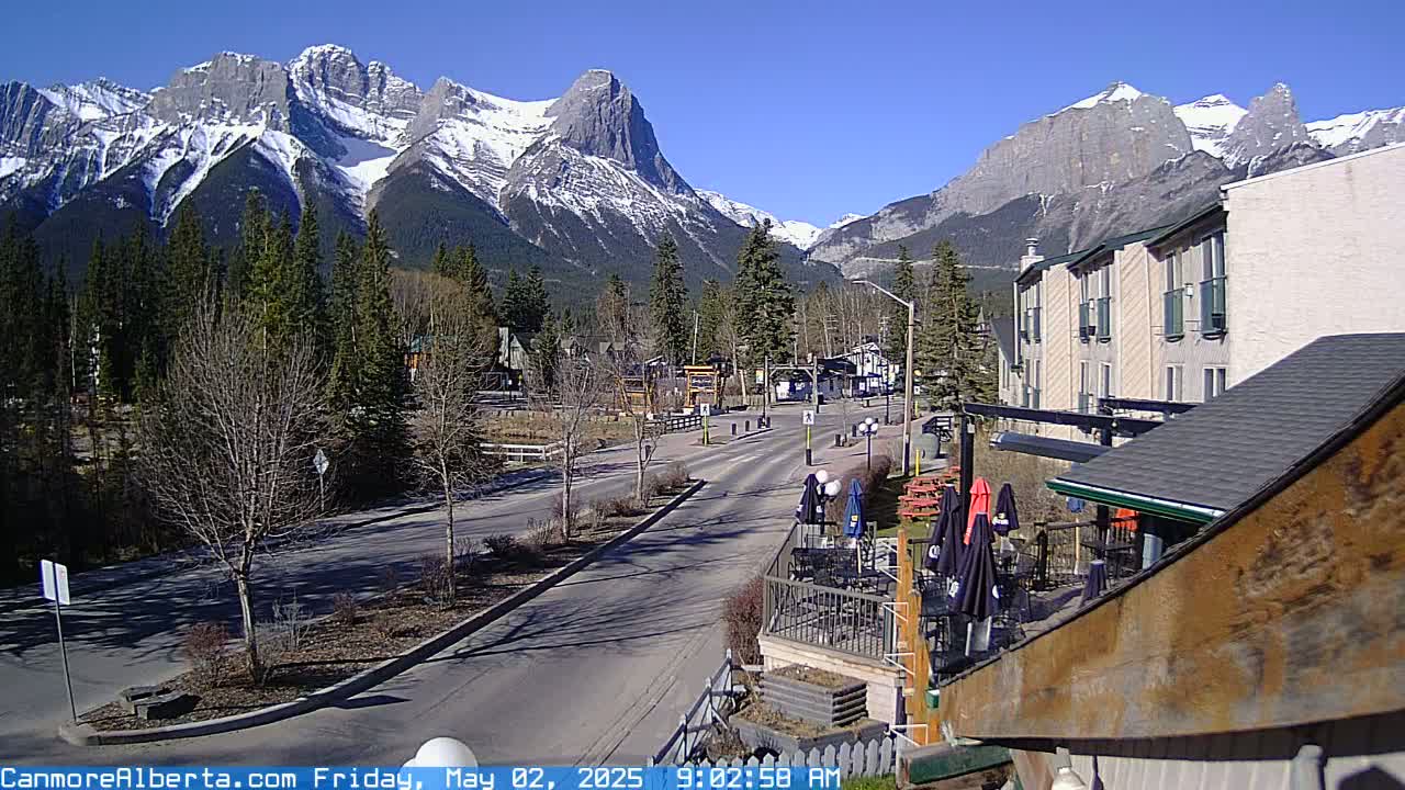 A sunny day reveals a mountain range capped with snow behind a quiet, tree-lined street with a few buildings and an outdoor patio.