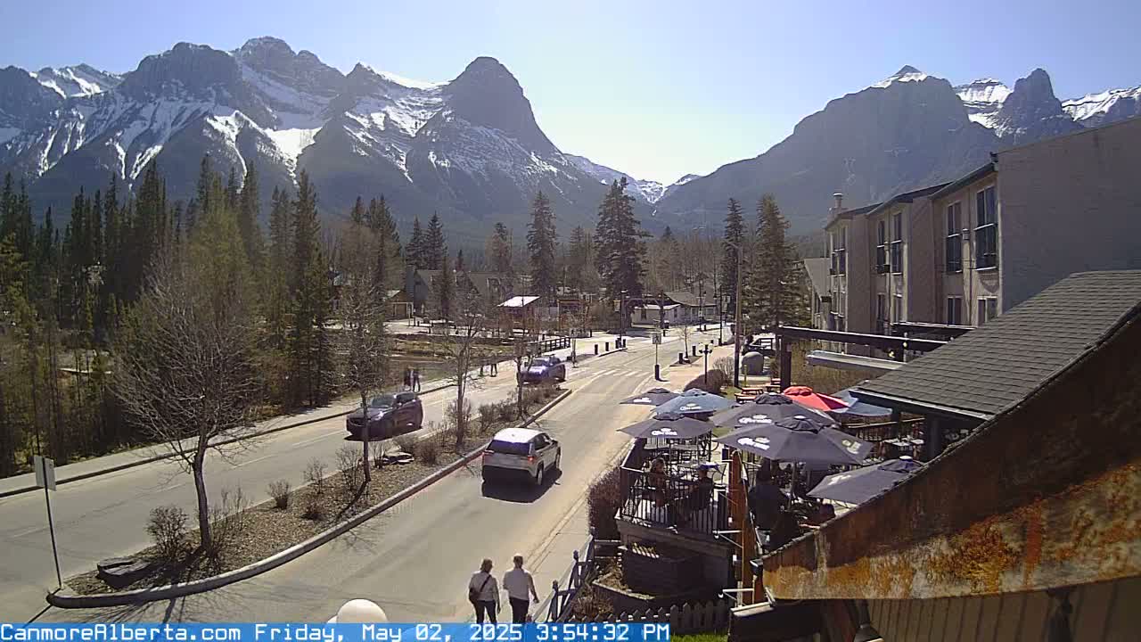 A sunny mountain town street scene shows cars driving on a paved road that leads towards snow-capped mountains, with buildings, trees, and outdoor seating visible.