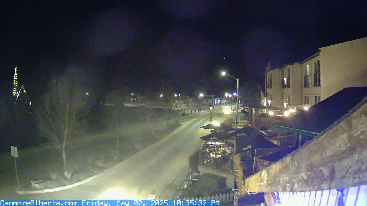 A snow-covered street at night, illuminated by streetlights, leads to a multi-story building with outdoor seating areas.