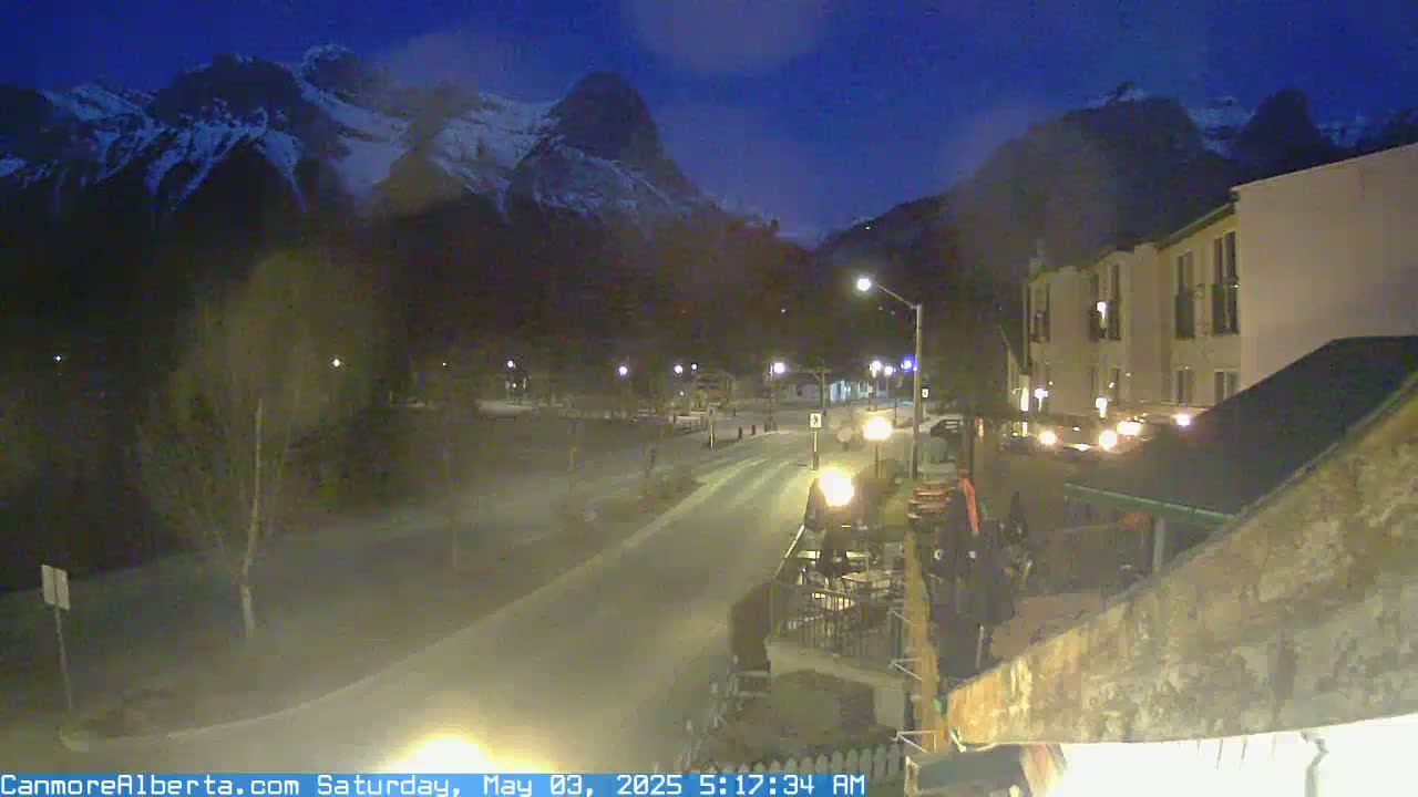 A dimly lit street scene at night shows snow-capped mountains in the background, with a road leading towards buildings under a dark, clear sky.
