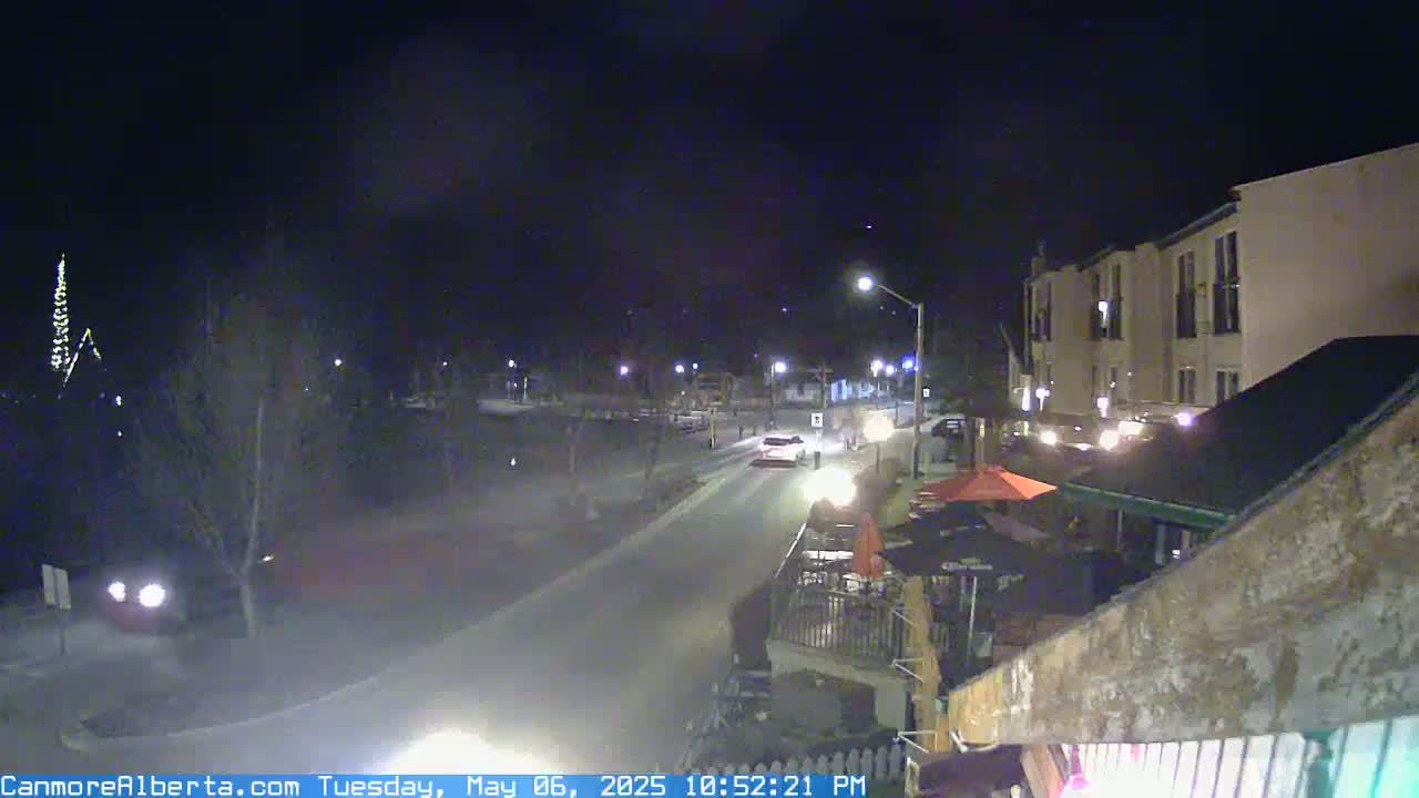 A nighttime street scene shows a few vehicles driving on a road past a building with outdoor seating under a clear, dark sky.