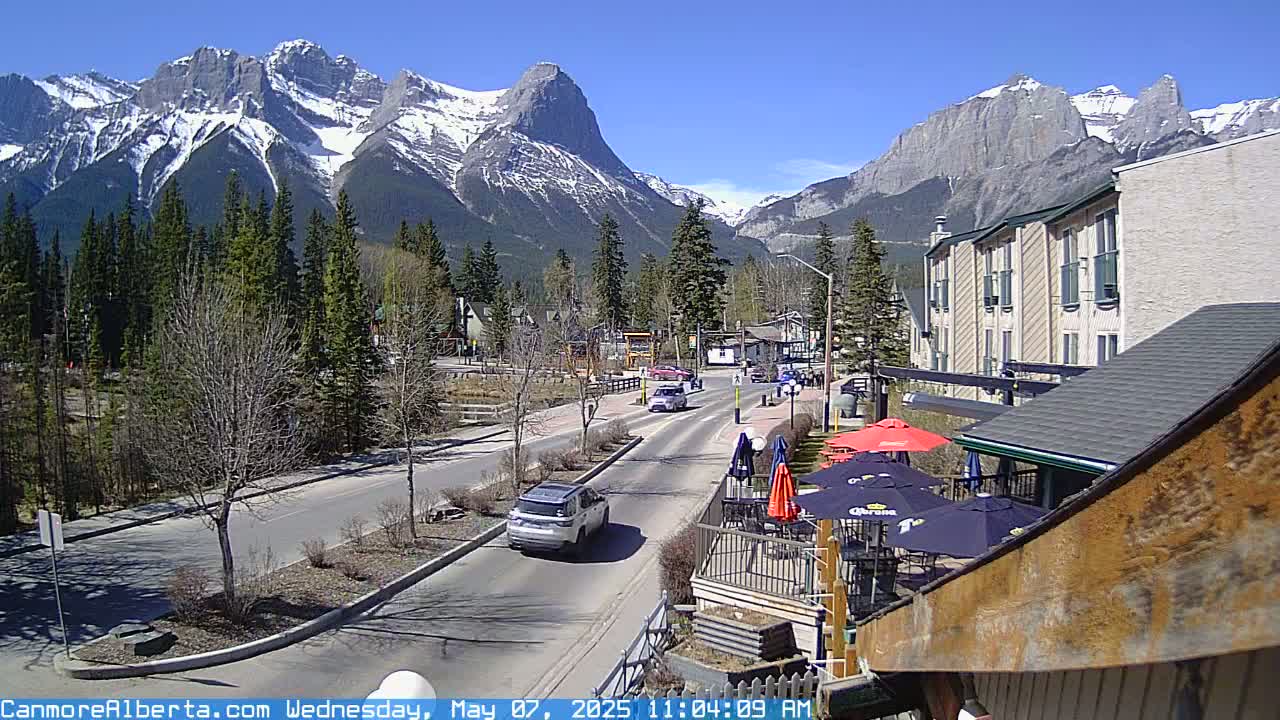 A sunny day reveals a mountain range capped with snow behind a town's main street, where cars drive past buildings and an outdoor patio with umbrellas.