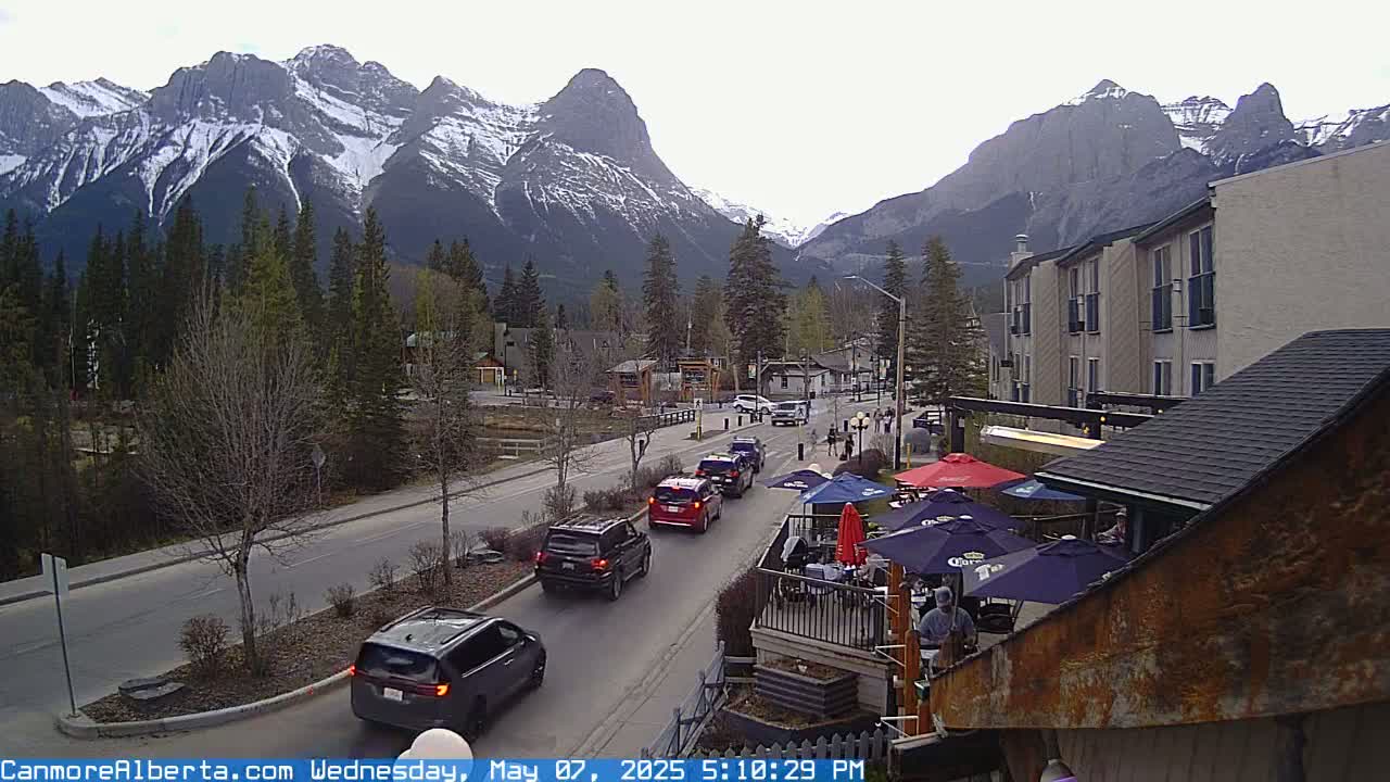 A mountain town street scene with overcast skies shows several vehicles driving on a road toward a snow-capped mountain range, and a patio with umbrellas sits in front of a building.