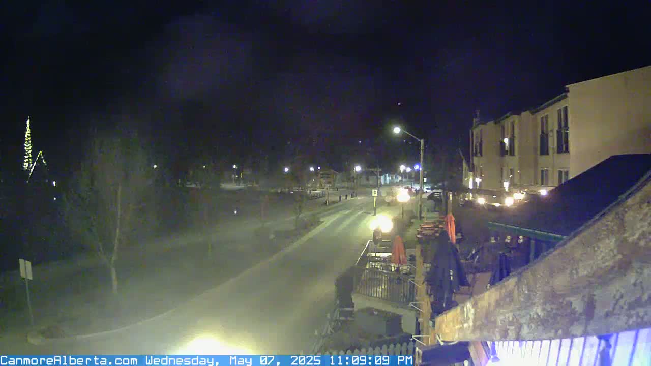 A nighttime view of a mostly empty street in front of a multi-story building, with some outdoor seating areas visible and a few cars parked.