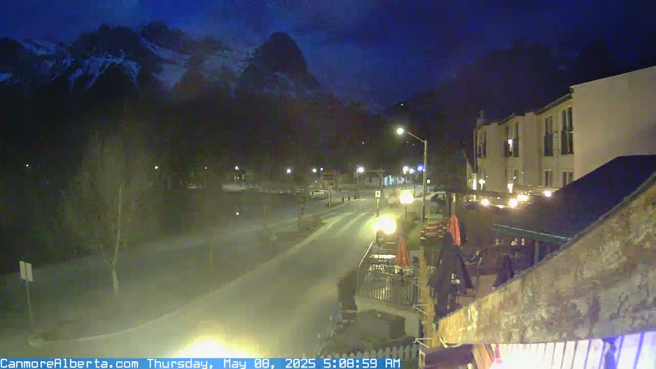 A nighttime view of a town street, illuminated by streetlights, with snow-capped mountains in the background under a dark blue sky.