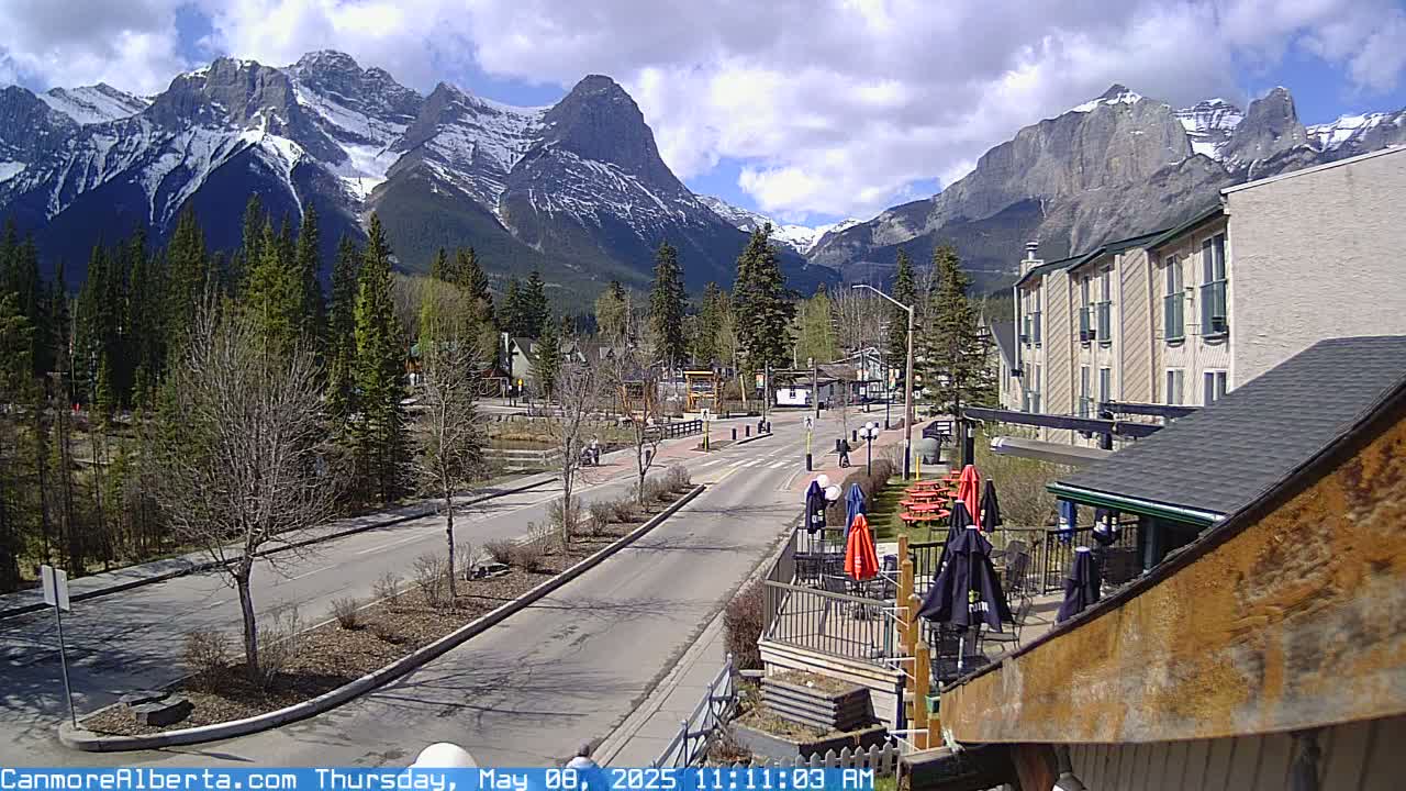A partly cloudy day reveals a mountain range partially covered in snow behind a quiet town street with a few pedestrians and outdoor patio seating.