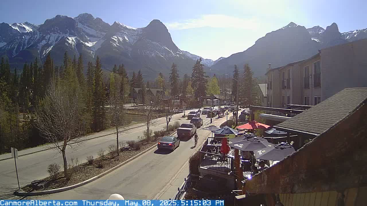 A sunny mountain town scene shows a street with cars, a cyclist, and an outdoor patio area in front of a building, all set against a backdrop of snow-capped mountains and evergreen trees.