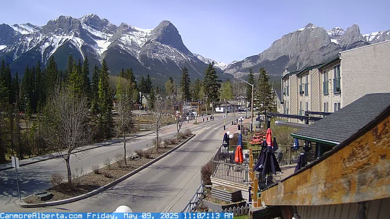 A paved road leads toward snow-capped mountains under a clear blue sky, with a town and evergreen trees in the foreground.