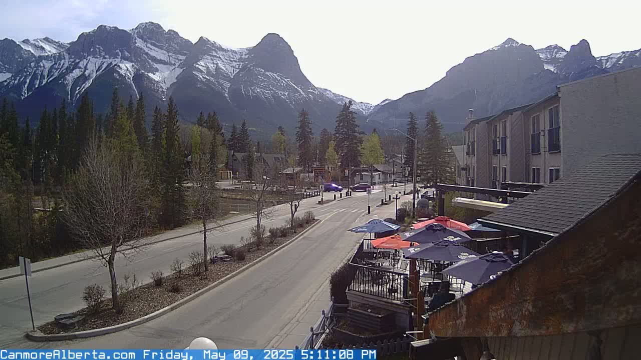 A mostly sunny day reveals a town nestled at the base of snow-capped mountains, with a street view showing a few cars and an outdoor patio area.
