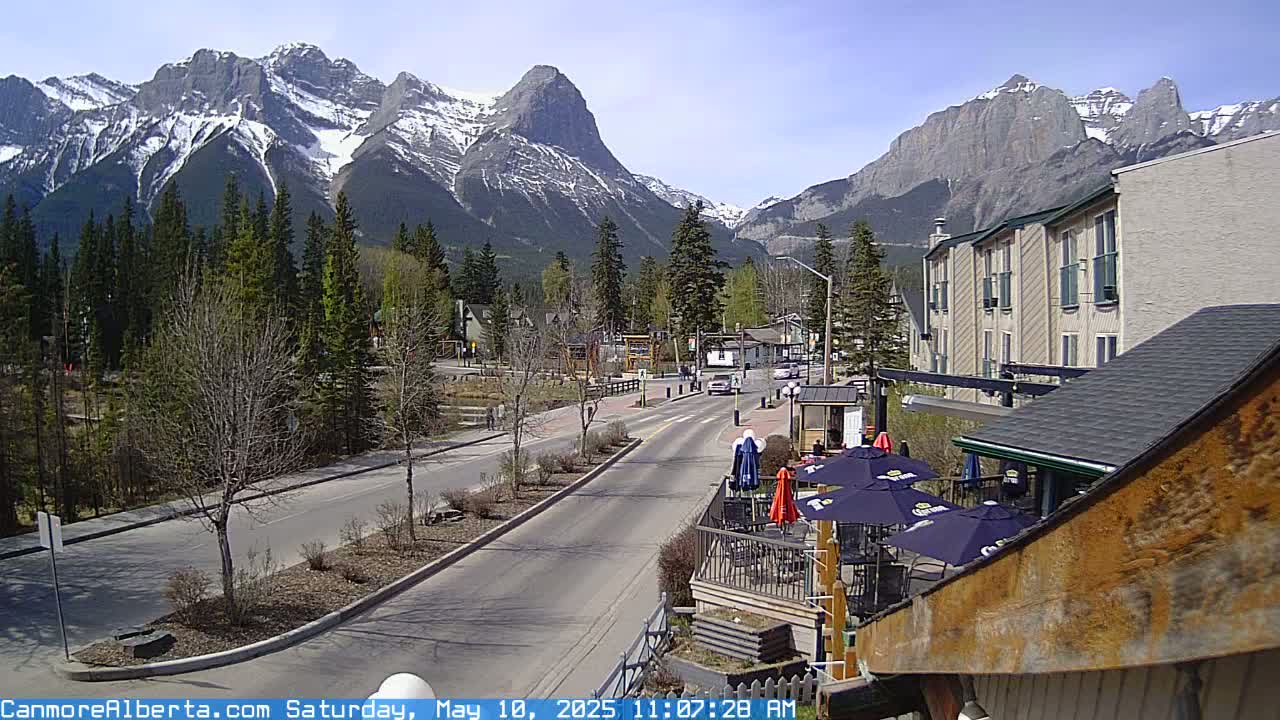 A sunny mountain town scene features a road with sparse traffic, lined with trees and buildings, with snow-capped mountains in the background.