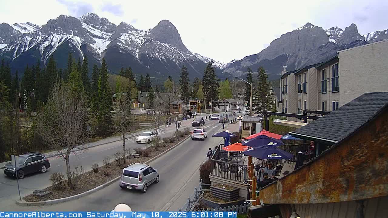 A partly cloudy day reveals a mountain range capped with snow, overlooking a town with cars driving on a street, and an outdoor patio area with people seated at tables under umbrellas.