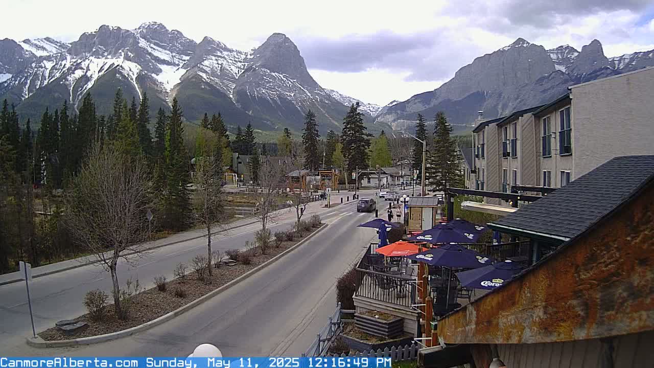 A partly cloudy day shows a mountain town street scene with snow-capped mountains in the background and outdoor patio seating at a restaurant in the foreground.