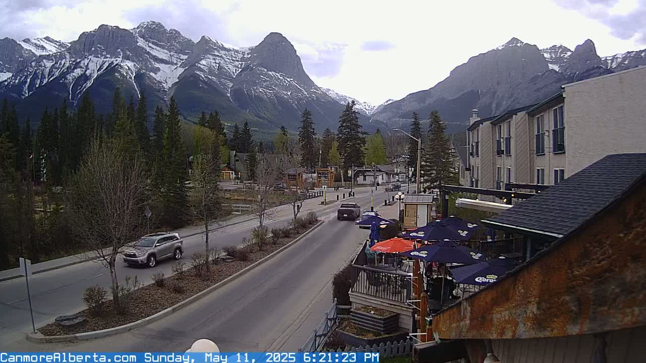 A partly cloudy day reveals a mountain town scene with snow-capped mountains in the background, a road with vehicles, and an outdoor patio area with umbrellas.