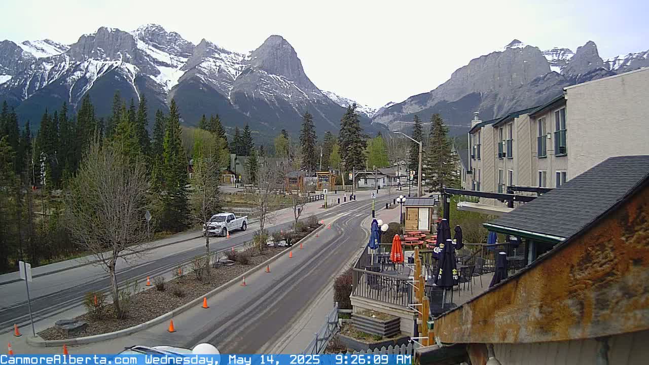 A partly cloudy day shows a mountain town with a road curving through it, leading towards snow-capped mountains in the background.