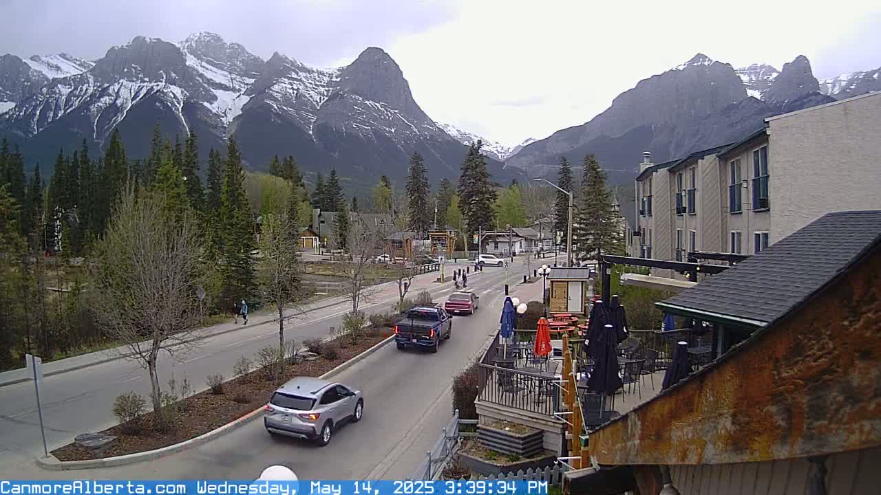 A partly cloudy day shows a mountain range with snow-capped peaks behind a town with cars on a road and an outdoor patio area.