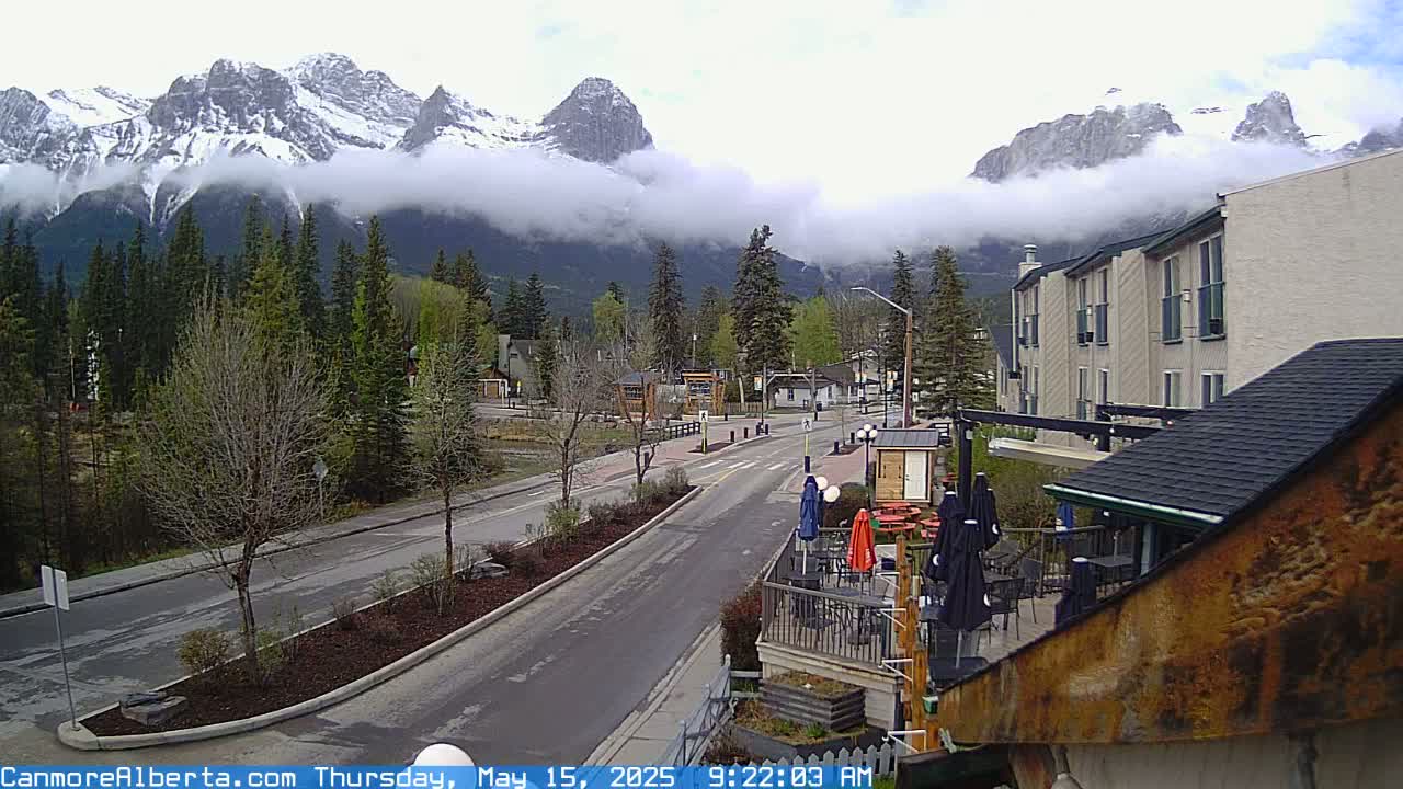 A partly cloudy day reveals snow-capped mountains shrouded in fog, overlooking a town with a street, buildings, and patio seating.
