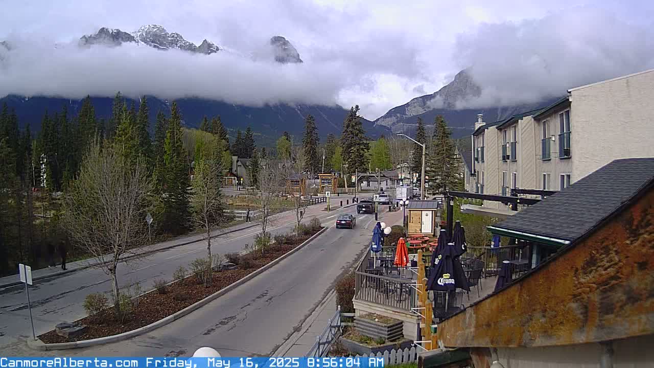 A mostly cloudy day reveals a mountain range partially obscured by low clouds above a town with a road and several buildings, including a multi-story hotel.