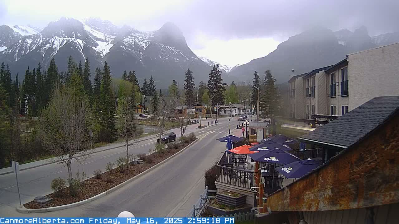 A town street scene shows snow-capped mountains under an overcast sky, with cars on the road and outdoor seating at a restaurant in the foreground.