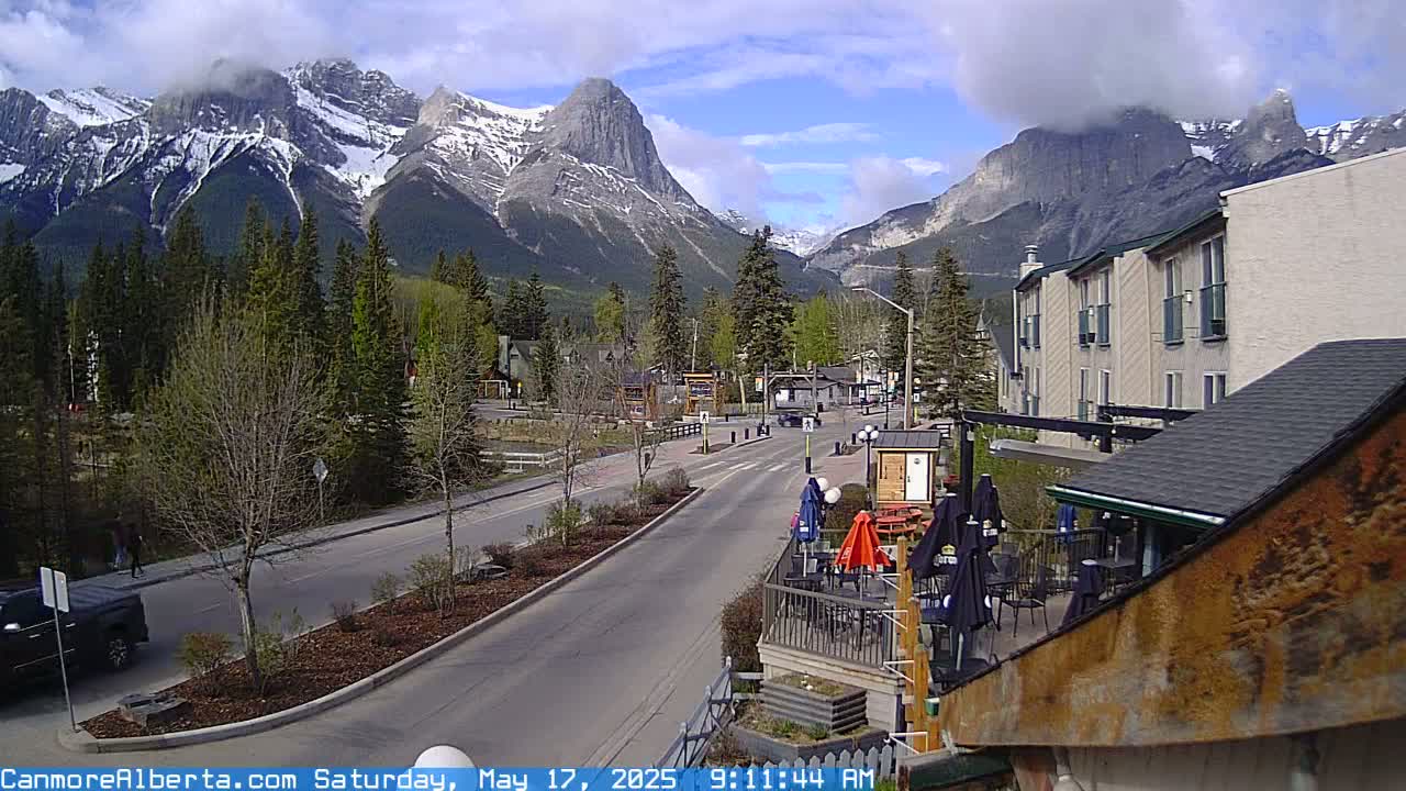 A partly cloudy day reveals a mountain range with snow-capped peaks overlooking a town with a street and buildings, including a patio with outdoor seating.