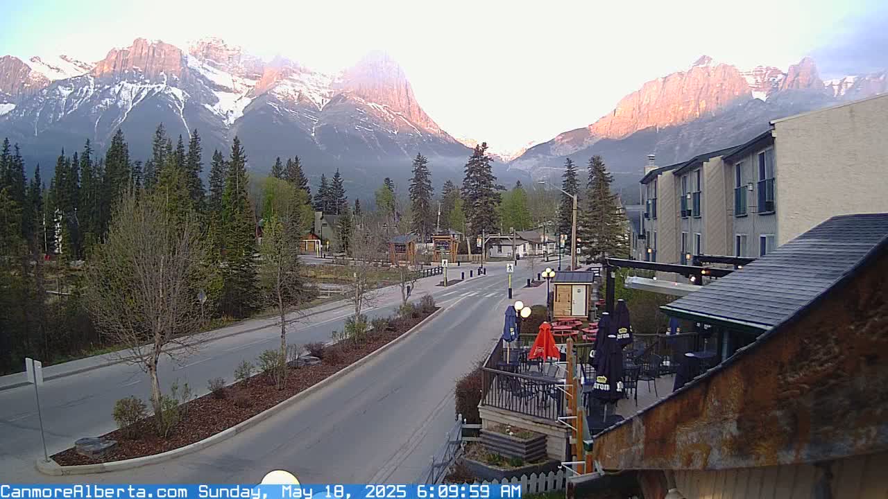 A mountain range at sunrise is visible beyond a town's street, which features buildings, landscaping, and outdoor seating.