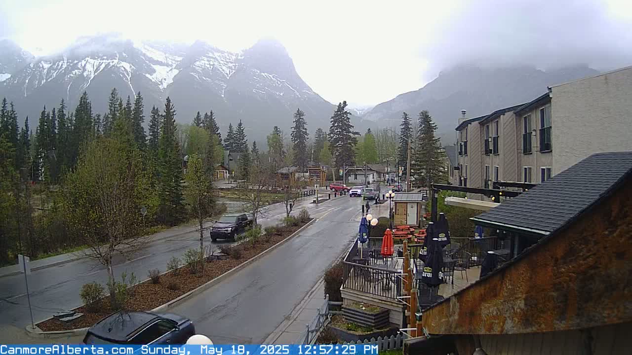 A wet, overcast day reveals a mountain town street scene with vehicles, buildings, and snow-capped mountains in the background.