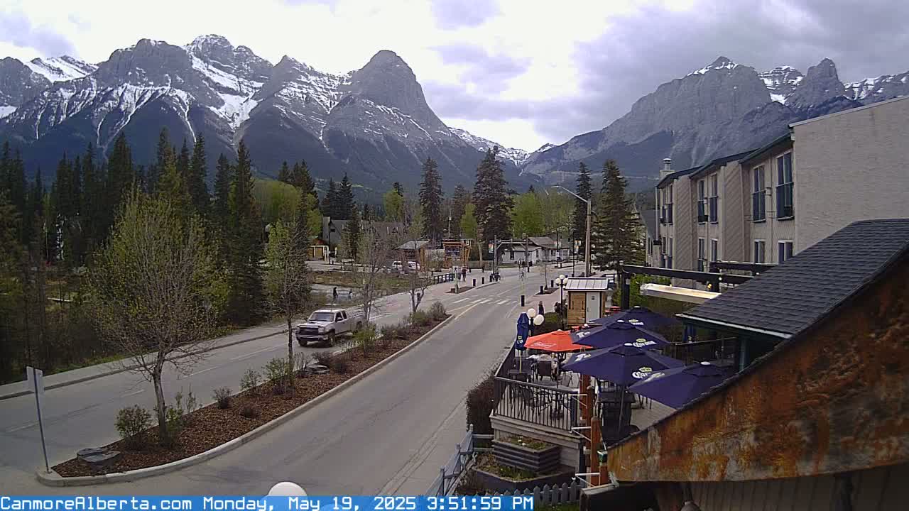 A partly cloudy day reveals a mountain range behind a town's main street with cars driving by and outdoor seating at a restaurant.