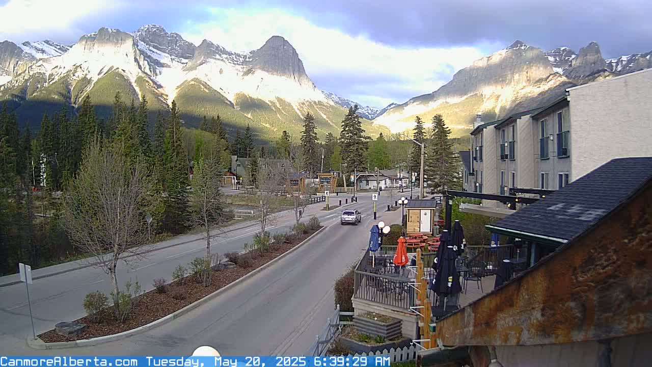A partly cloudy morning reveals a mountain town nestled at the base of snow-capped peaks, with a road leading toward them and an outdoor patio area visible in the foreground.
