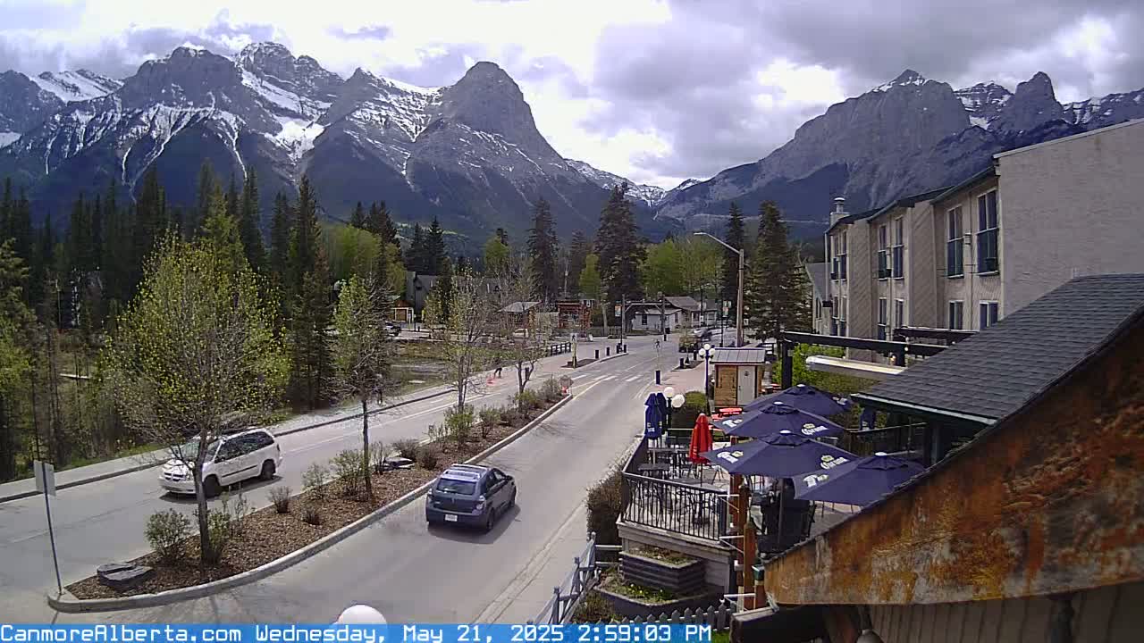 A partly cloudy day shows a mountain range behind a town with a street, cars, and outdoor patio seating.