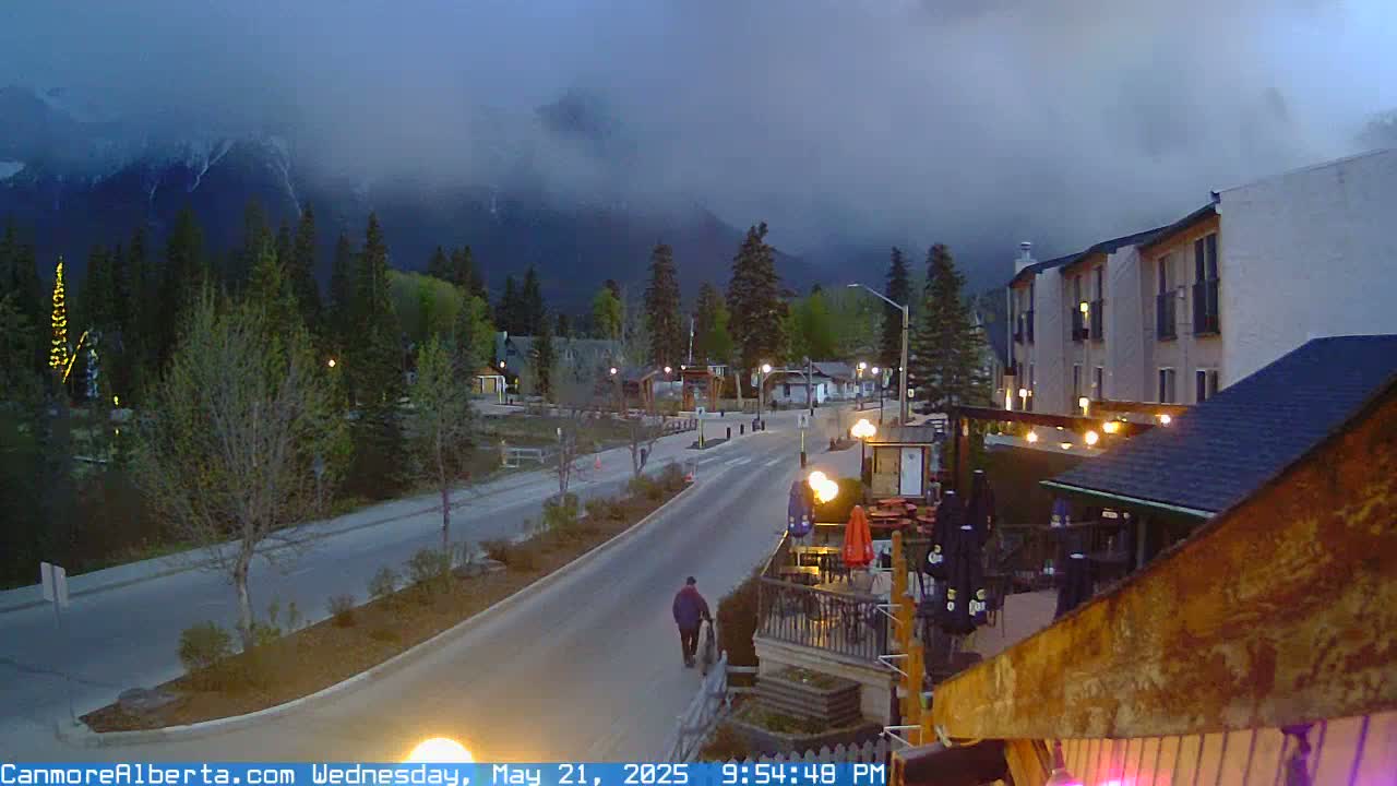 A mostly cloudy twilight scene shows a road winding through a town nestled at the base of a mountain range, with a person walking along the roadside.