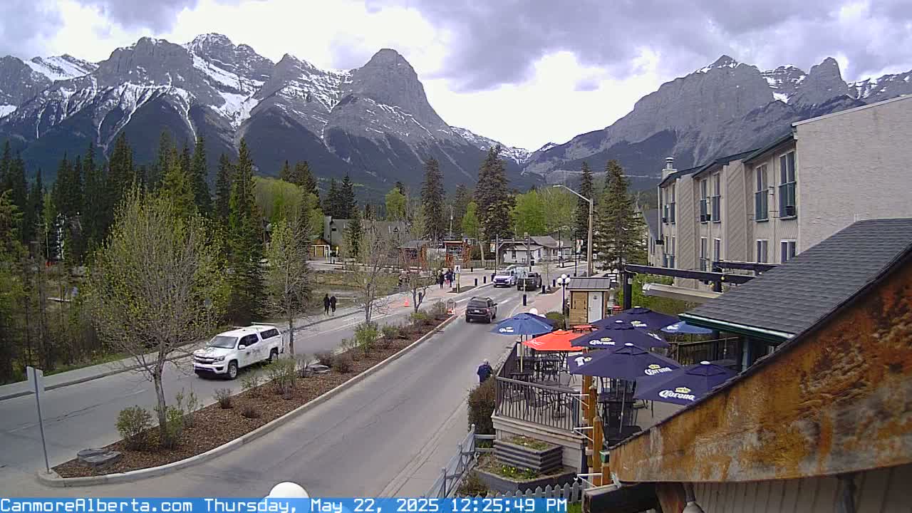 A street scene shows a partly cloudy day with snow-capped mountains in the background, vehicles on a road passing by a restaurant patio with umbrellas.