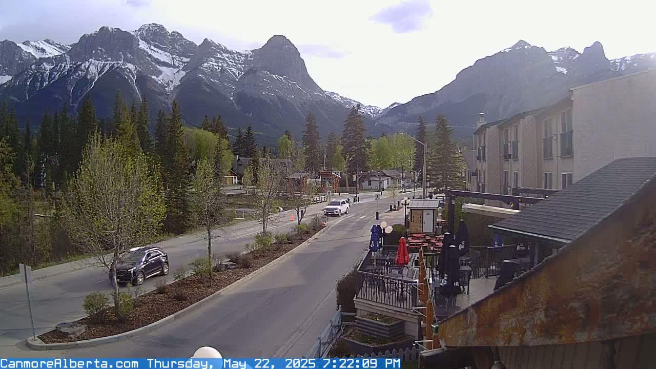 A partly cloudy day reveals a mountain range behind a town with cars on the road and an outdoor patio area of a building in the foreground.