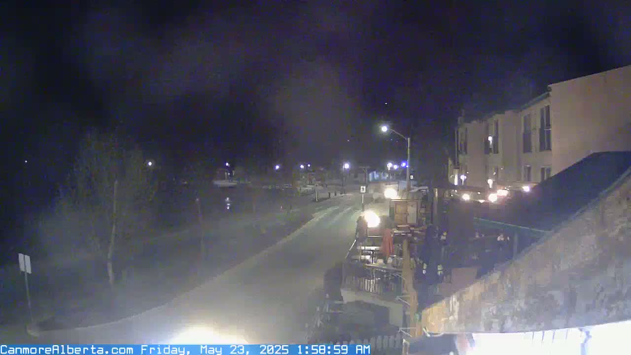A nighttime, slightly hazy view of a street scene with a multi-story building and a patio area, showing some outdoor lighting and a few vehicles.