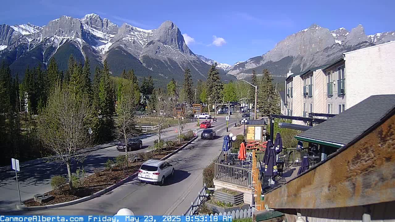 A sunny mountain town street scene shows several cars driving on a road lined with trees and buildings, with a mountain range in the background.