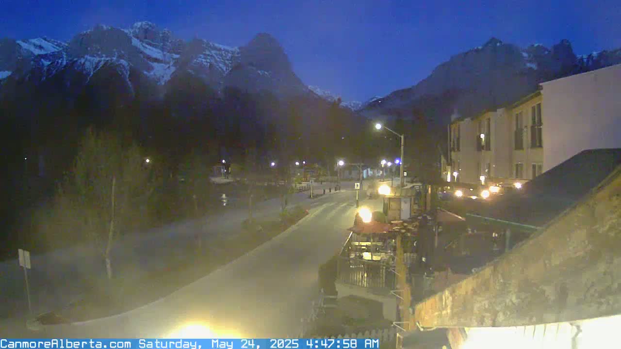 A nighttime, dimly lit street scene shows a road curving past buildings and toward snow-capped mountains under a dark blue sky.