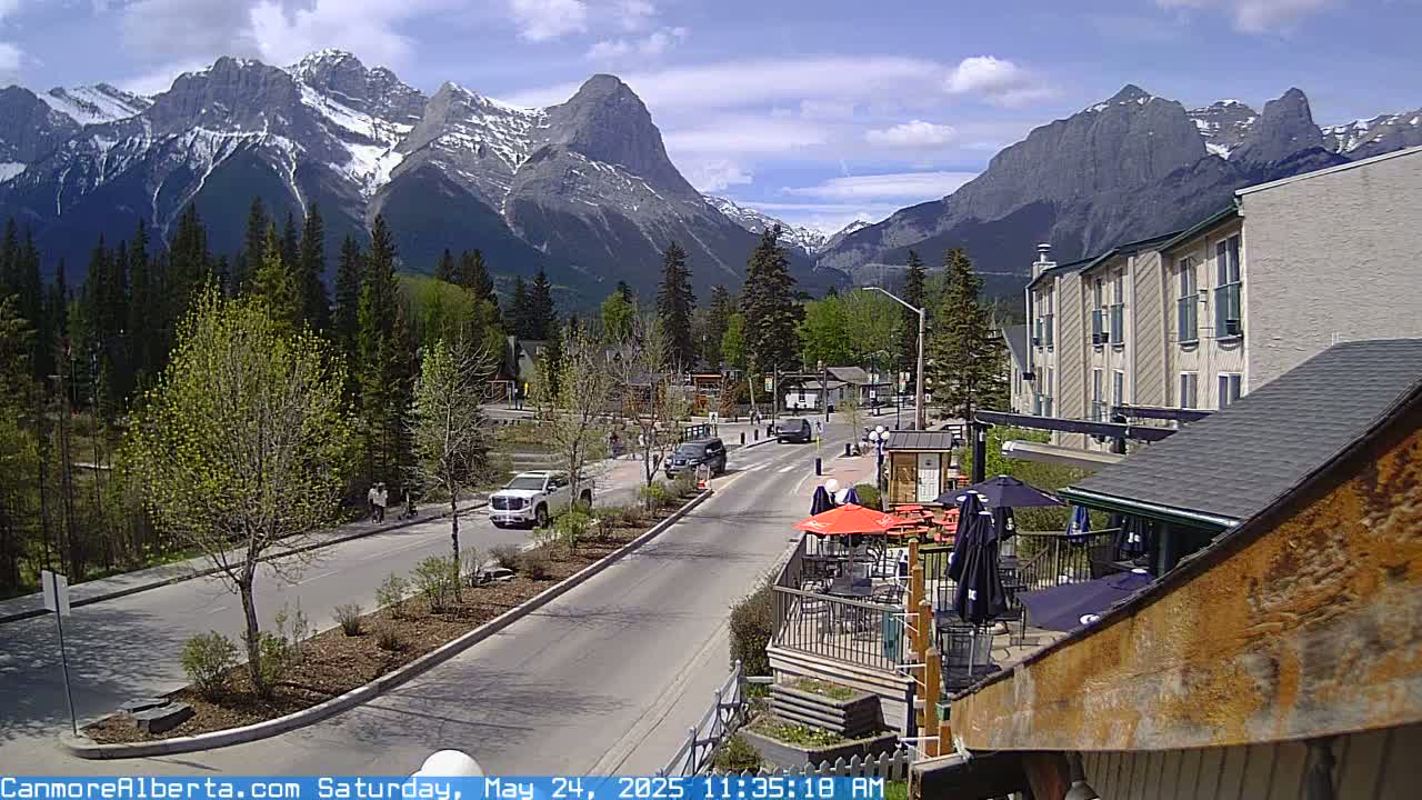 A partly sunny day reveals a mountain town scene with a road, vehicles, pedestrians, and buildings, all set against a backdrop of snow-capped mountains and green trees.
