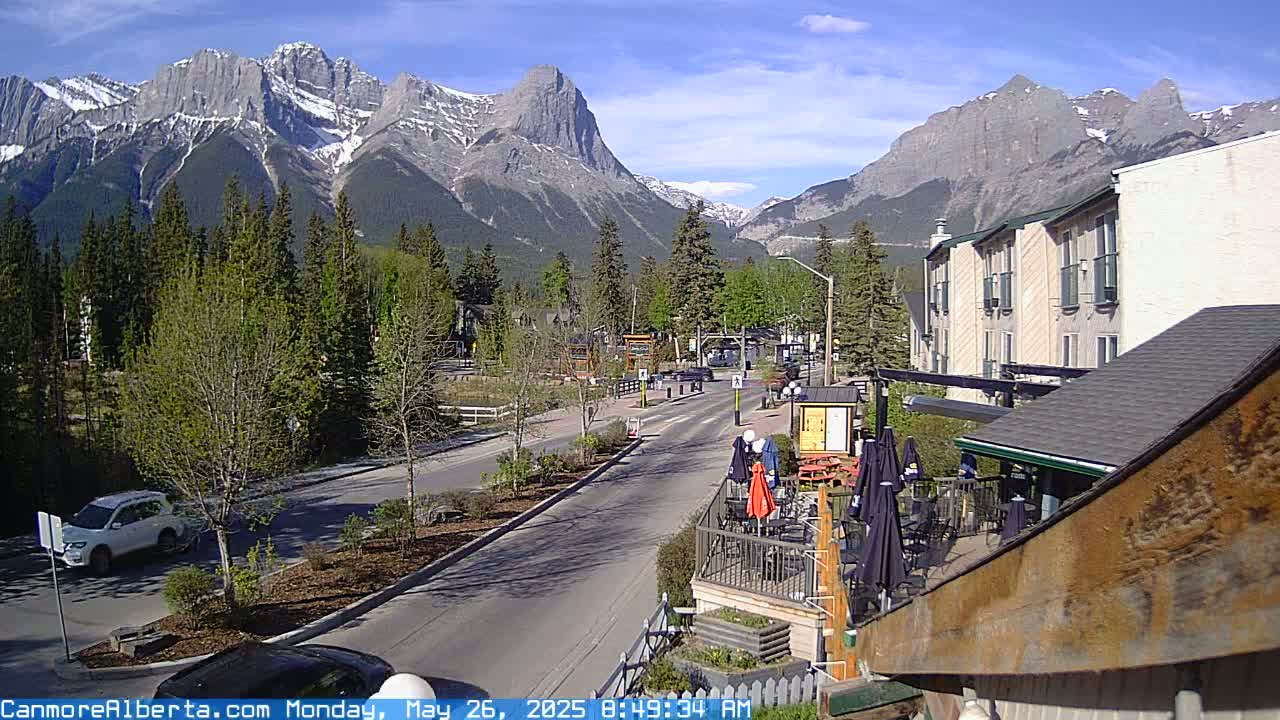 A sunny day in a mountain town shows a street scene with cars, trees, buildings, and snow-capped mountains in the background.