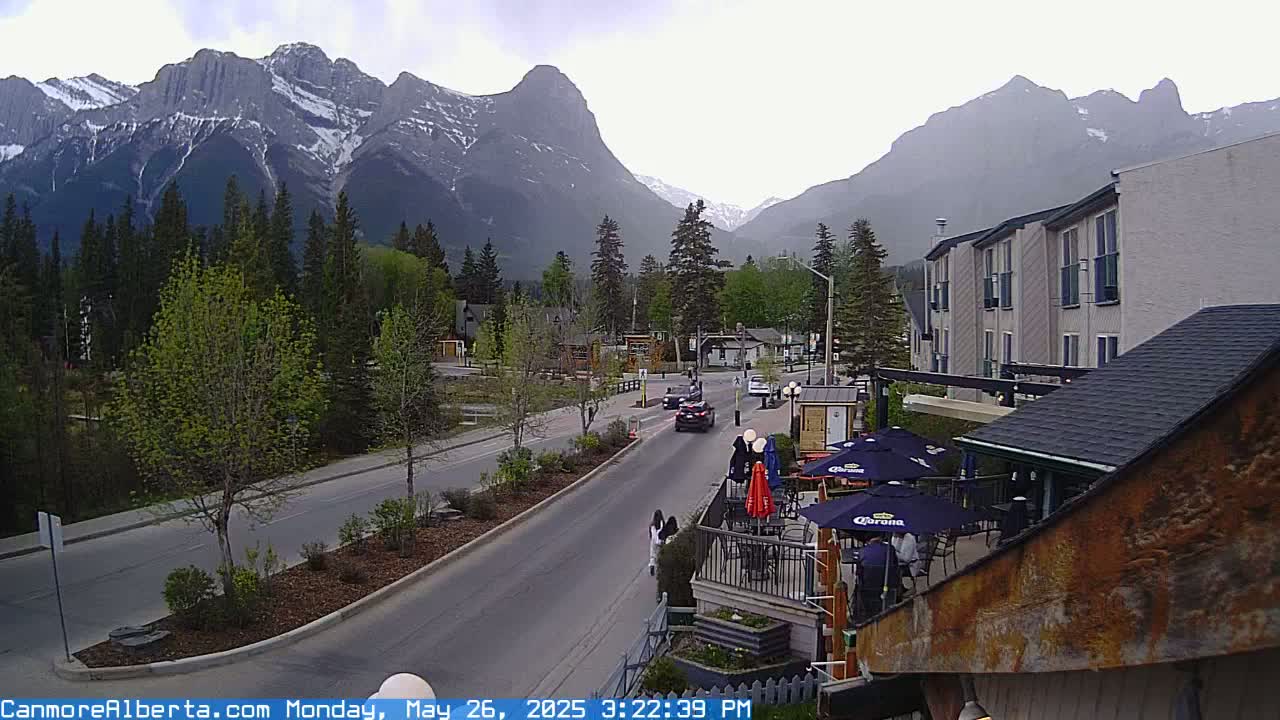 A street scene shows a road with cars, pedestrians, and outdoor seating at a restaurant, all set against a backdrop of snow-capped mountains under an overcast sky.