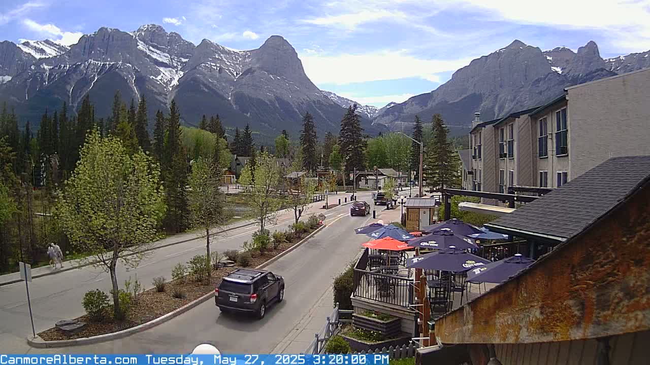 A mountain town street scene on a partly sunny day shows a roadway with cars, a pedestrian, trees, buildings, and outdoor seating with umbrellas beneath a backdrop of snow-capped mountains.