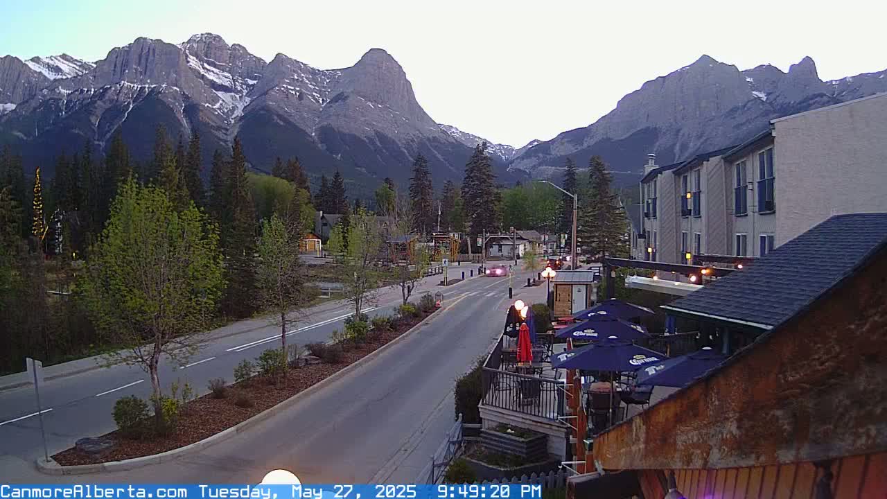 A mountain town street scene at dusk shows a road with cars, a few buildings, and trees, all set against a backdrop of majestic, snow-capped mountains.