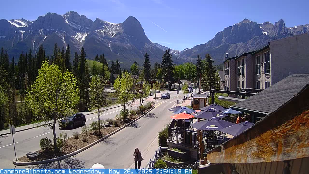 A sunny mountain town street scene features a road with cars, outdoor patio seating, and a multi-story building, all set against a backdrop of snow-capped mountains.