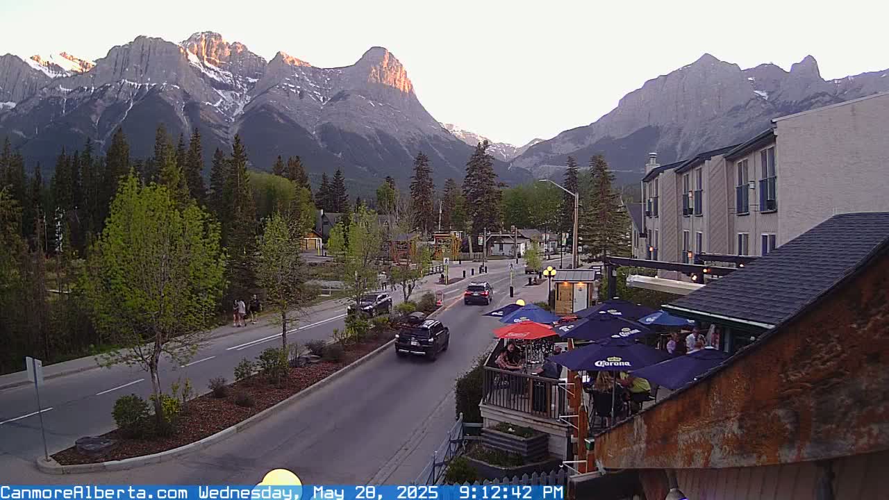 A mountain town street scene at sunset shows cars driving on a road lined with trees and buildings, with a mountain range in the background.