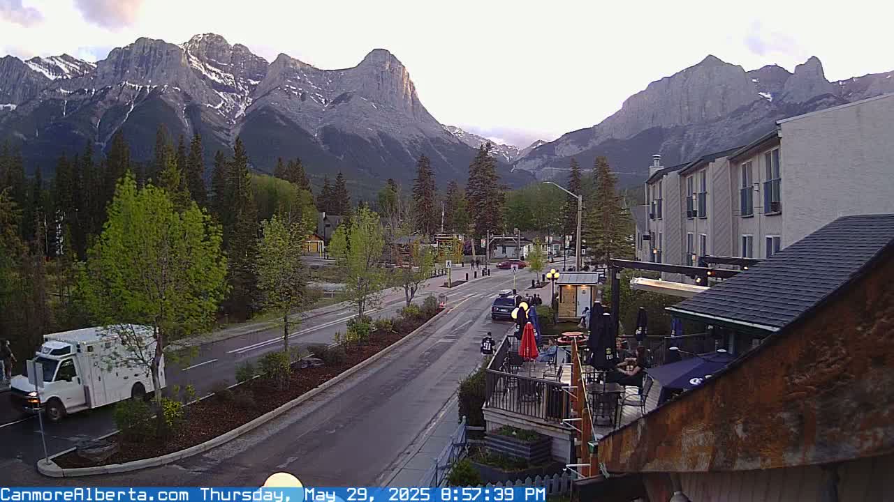 A mountain town street scene at dusk, shows a road with some vehicles, trees, buildings, and a patio area, all set against a backdrop of imposing mountains.