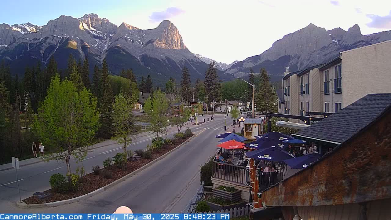 A mountain town street scene at dusk, featuring a road leading towards imposing mountains under a partly cloudy sky, with several buildings and people visible.