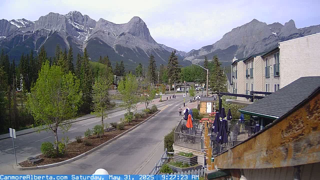 A partly sunny day reveals a mountain range behind a town with a paved road, trees, and a building with an outdoor patio.